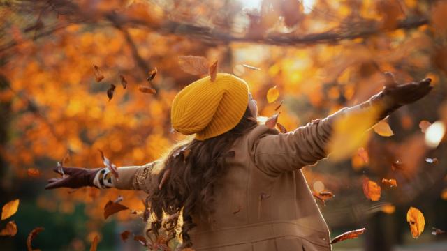 Seen from behind middle aged woman in beige coat and orange hat rejoicing outdoors on the city park in autumn.
