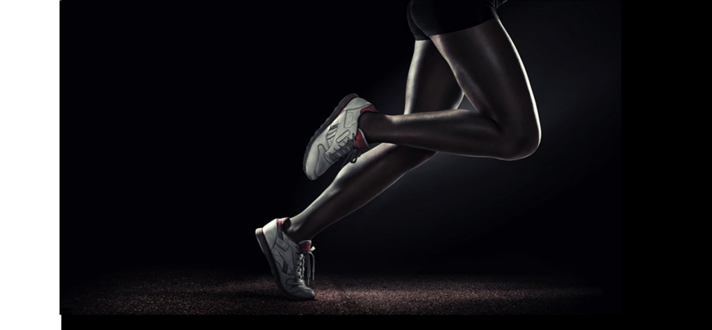 Photograph of runner’s legs mid-stride in gray shoes and black leggings on dark background