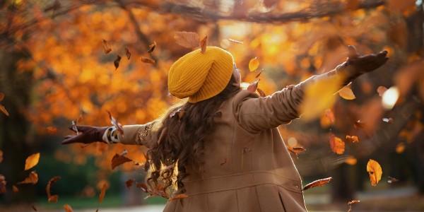 Seen from behind middle aged woman in beige coat and orange hat rejoicing outdoors on the city park in autumn.