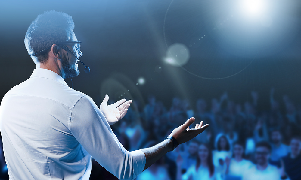 A speaker in a white shirt gestures towards an audience under bright stage lights.