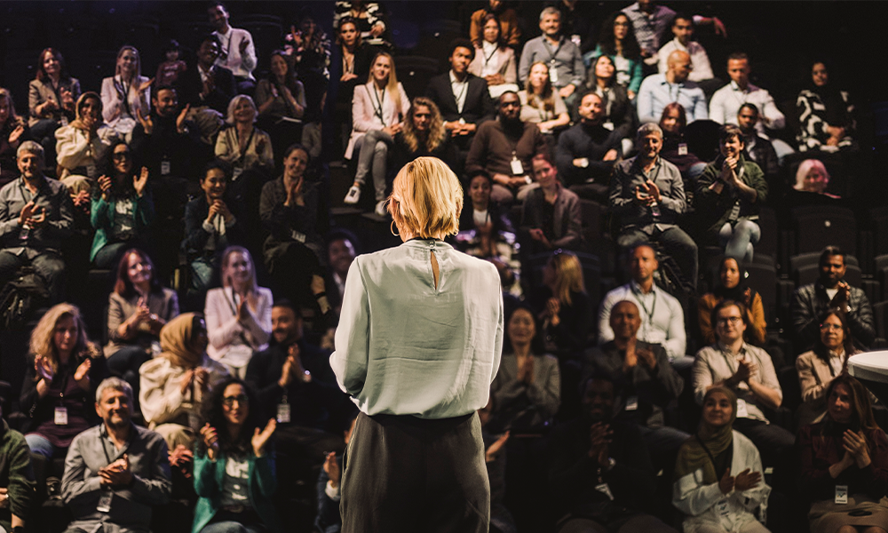 A person stands on stage, facing an applauding audience in a theater setting.