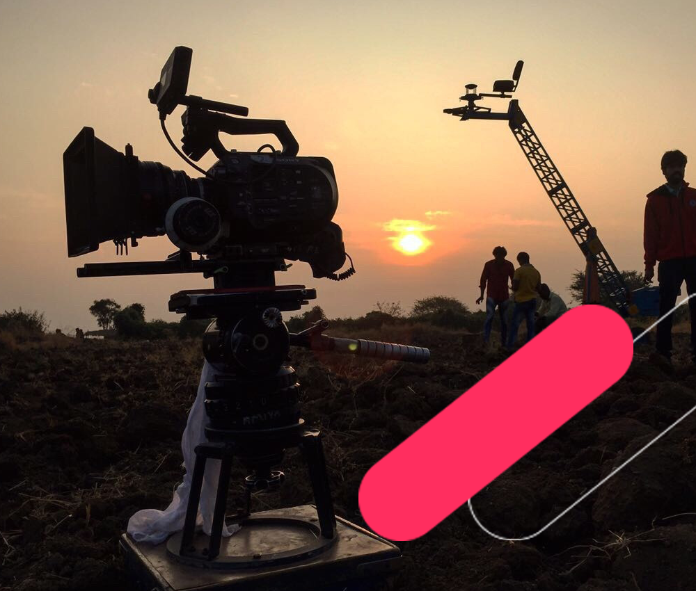 Film camera on tripod silhouetted against sunset with crane and crew on outdoor set